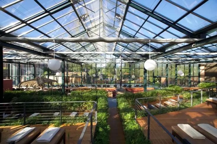 Innenraum eines großen Glasgewächshauses mit Glasdach und Metallrahmen, hängenden Kugelleuchten, mehreren Pflanzbeeten, Holzstegen, Sitzbänken und zentralem Fußweg