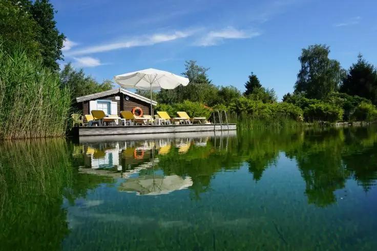 Kleines Holzhaus am Ufer mit Steg, weißem Sonnenschirm, gelben Liegestühlen, Rettungsring, umgeben von Bäumen und Schilf, Spiegelung im ruhigen Wasser