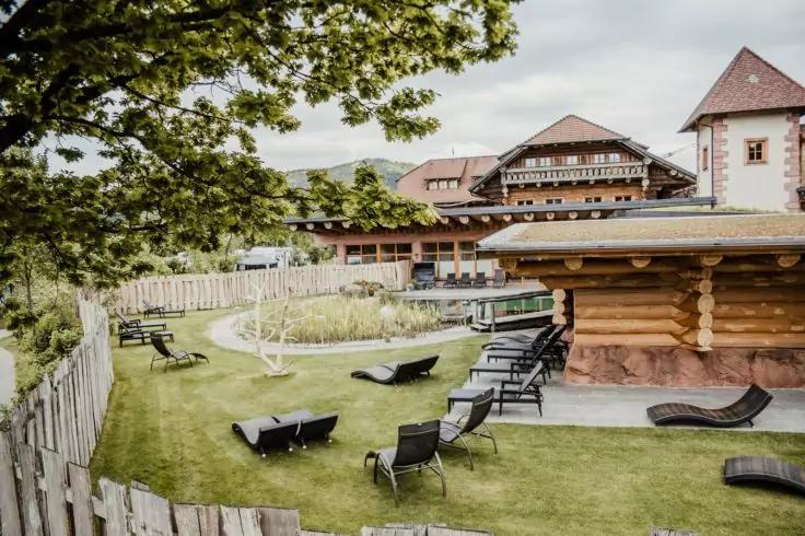 Rasenfläche mit mehreren Liegestühlen vor einem Holzhaus und größeren Gebäuden, Holzzaun links, Baum im Vordergrund, bewölkter Himmel