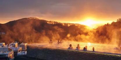 Au&szlig;enbecken mit dampfendem Wasser und mehreren Personen im Wasser, Berge im Hintergrund, Sonne am Horizont, orangefarbener Himmel