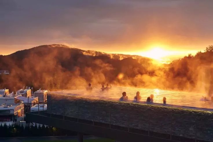 Au&szlig;enbecken mit dampfendem Wasser und mehreren Personen im Wasser, Berge im Hintergrund, Sonne am Horizont, orangefarbener Himmel