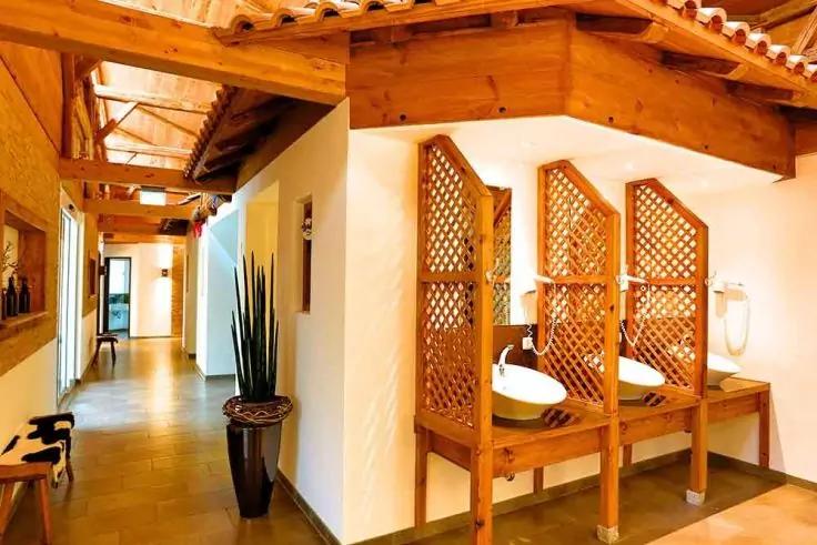 Zwei weiße Aufsatzwaschbecken auf einer Holzablage, dahinter dreiteilige Holz-Gittertrennwände; rechts ein wandmontiertes Gerät; Holzdeckenbalken, gefliester Boden, hoher Pflanzenkübel im Flur und Tür am Ende