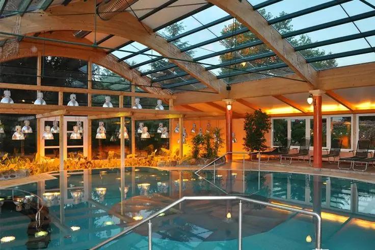 Hallenbad mit Glasdach und Holzträgern, Treppen mit Edelstahlgeländern, Pflanzen, Liegestühle und warmes Licht