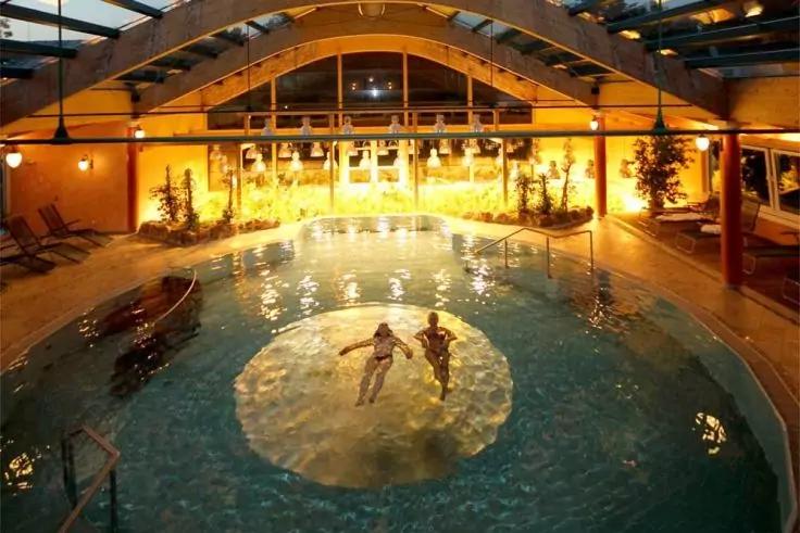 Innenpool mit rundem, flachem Bereich in der Mitte; zwei Personen im Wasser; gelbe Beleuchtung, Pflanzen, Liegestühle an den Seiten und geschwungene Deckenbalken