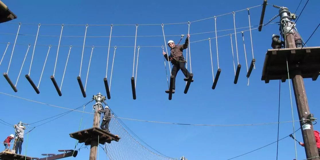 Mehrere Personen mit Helm und Sicherungsgurt auf schwingenden Holzstegen zwischen Plattformen und Pfosten vor blauem Himmel