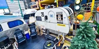 Wei&szlig;er kleiner Wohnwagen mit schwarzen Streifen und Porsche-Schriftzug, silberner Anh&auml;nger daneben, Reifen auf dem Boden, Regale mit Werkzeug und ein k&uuml;nstlicher Tannenbaum