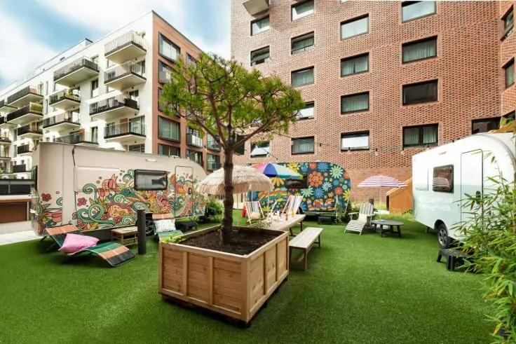 Innenhof mit k&uuml;nstlichem Rasen, zwei bunt bemalte Wohnwagen, rechteckiger Holzpflanzkasten mit Baum, Tische, St&uuml;hle, Sonnenschirme und umliegende Wohngeb&auml;ude