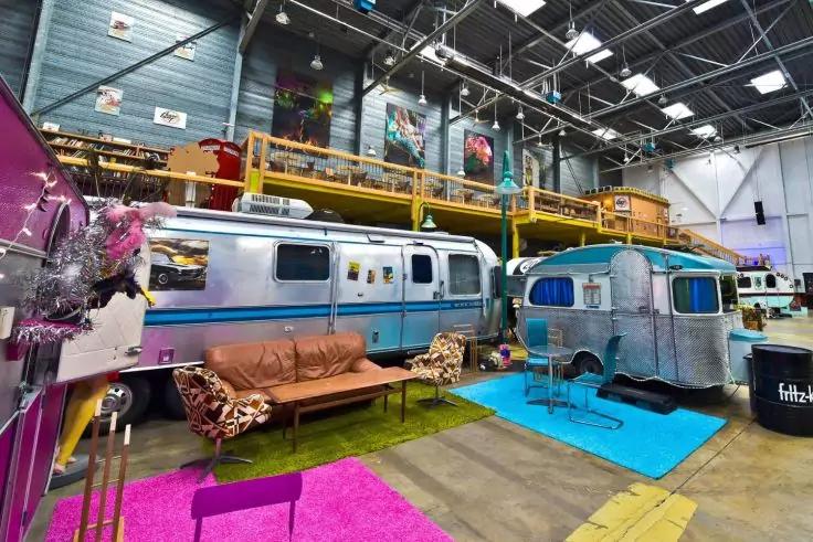 Innenraum einer gro&szlig;en Halle mit zwei silbernen Wohnwagen (Airstream), braunem Ledersofa, bunten Teppichen (pink, t&uuml;rkis, gr&uuml;n), gelber Empore mit Gel&auml;nder, Oberlichtern und Industriebeleuchtung