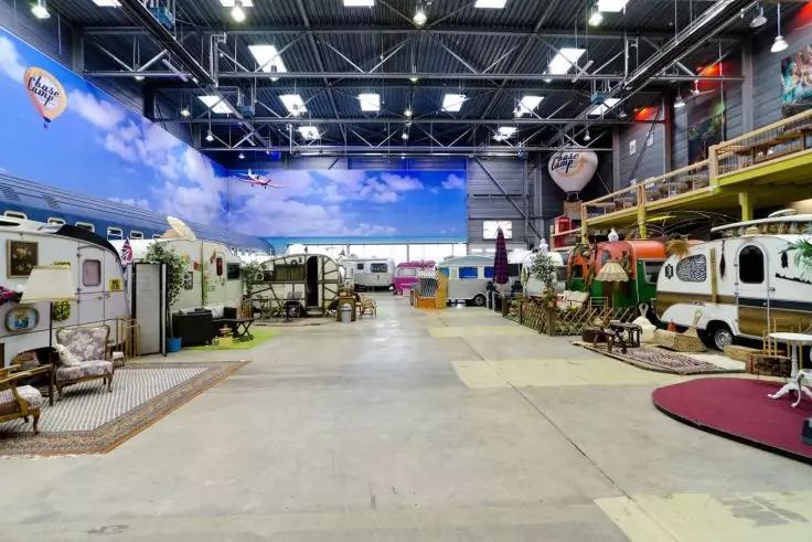 Gro&szlig;e Halle mit hohem Dach und Deckenbeleuchtung; links und rechts Reihen von Wohnwagen und Anh&auml;ngern, davor M&ouml;bel, Teppiche und Dekoration; Betonboden; gro&szlig;e Wand mit Wolkenmotiv im Hintergrund