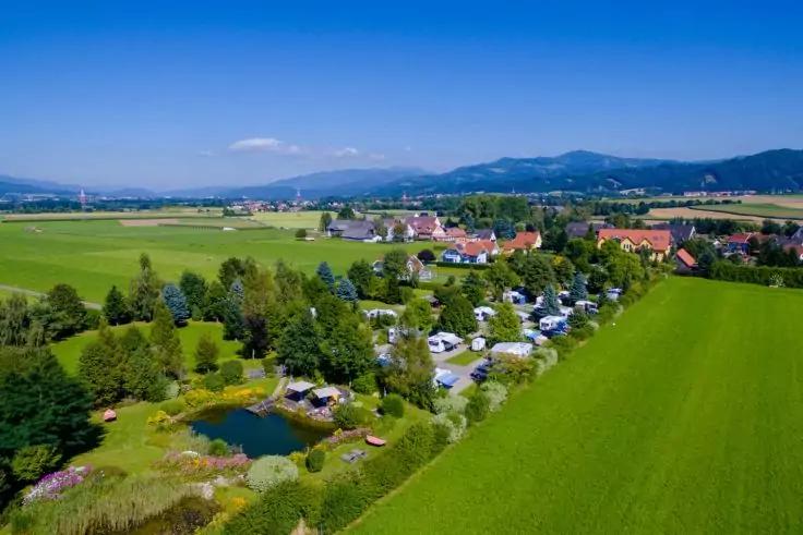 Luftaufnahme eines Dorfes mit H&auml;usern, gr&uuml;nen Feldern, B&auml;umen, einem Campingplatz mit Wohnmobilen und einem kleinen Teich, H&uuml;gel im Hintergrund unter blauem Himmel