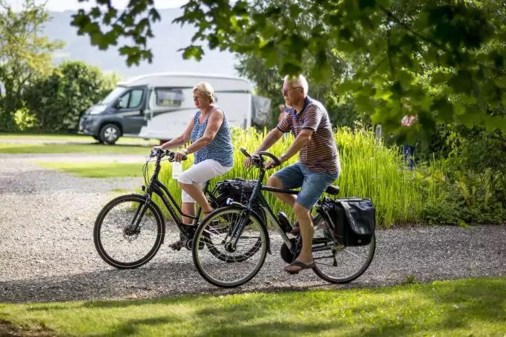 Zwei Personen fahren nebeneinander Fahrrad auf einem geschotterten Weg; im Hintergrund ein wei&szlig;es Wohnmobil und B&auml;ume