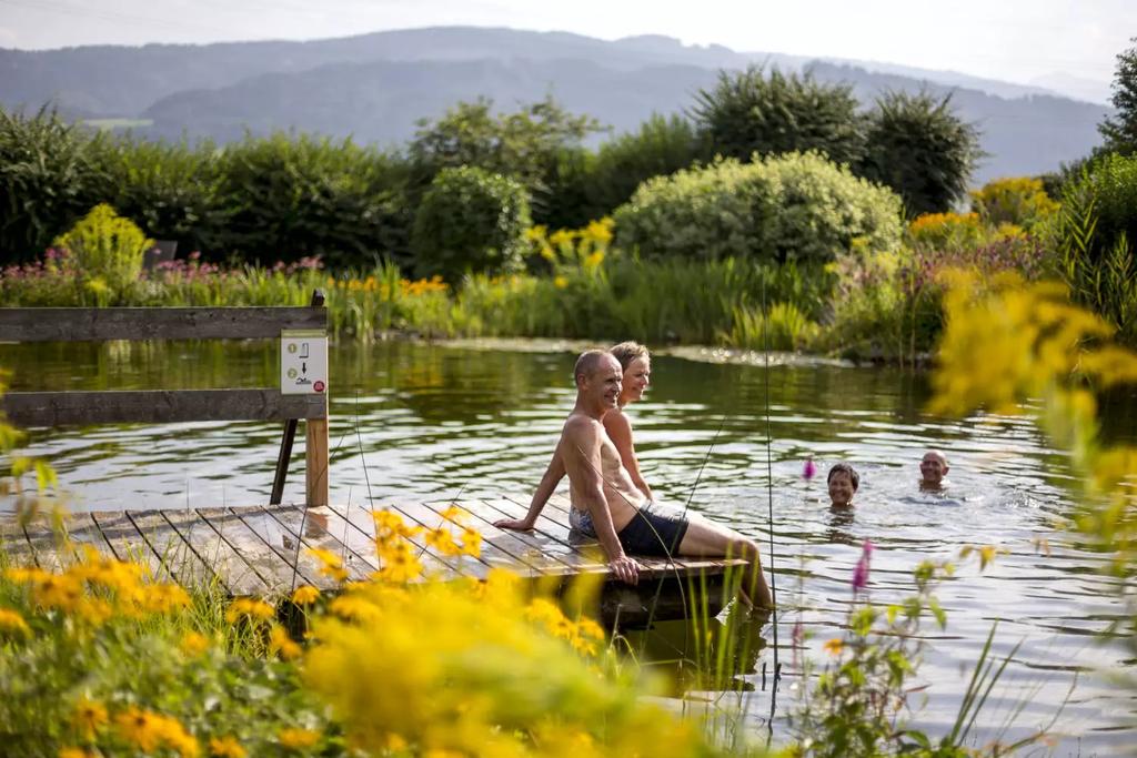 Zwei Personen in Badebekleidung sitzen auf einem Holzsteg an einem Teich. Mehrere Personen schwimmen im Wasser. Ufer mit gelben Blumen, B&uuml;schen und h&uuml;geliger Landschaft im Hintergrund