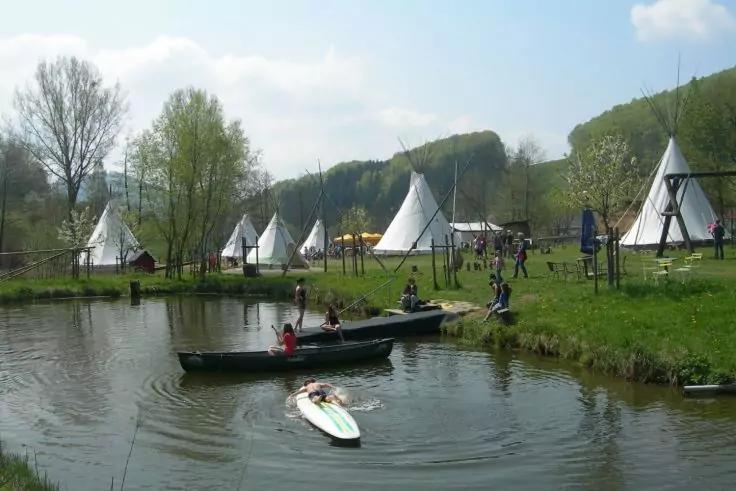 Teich mit Personen in kleinen Booten und auf einem Stand-up-Paddle, grasbewachsene Ufer, mehrere wei&szlig;e Tipis, B&auml;ume und H&uuml;gel im Hintergrund, blauer Himmel mit vereinzelten Wolken