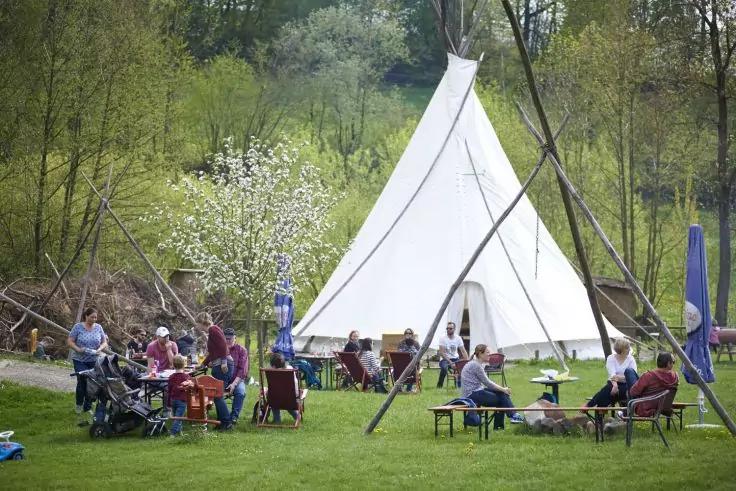 Wei&szlig;es Tipi auf gr&uuml;ner Wiese, Personen sitzen an Tischen, B&auml;nken und St&uuml;hlen, Kinderwagen sichtbar