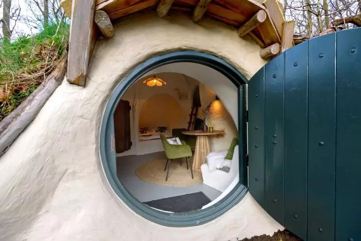 Runde Eingangst&uuml;r in einem Erdh&uuml;gel, gr&uuml;ne runde T&uuml;r ge&ouml;ffnet; Blick in einen geschwungenen, wei&szlig; verputzten Innenraum mit Holzdeckenbalken, rundem Holztisch, gr&uuml;ner und wei&szlig;er Stuhl, h&auml;ngender Lampe und Regalen