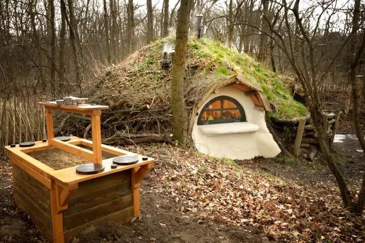 Kleines erdgedecktes Geb&auml;ude mit begr&uuml;ntem Dach und rundem Fenster; davor h&ouml;lzernes Hochbeet, Laub am Boden und kahle B&auml;ume