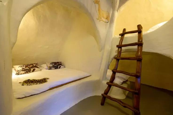 Wei&szlig; verputzter, gew&ouml;lbter Raum mit eingebauter Liegefl&auml;che, Matratze, gemusterten Kissen, h&ouml;lzerner Leiter an der Wand und warm gelbem Licht