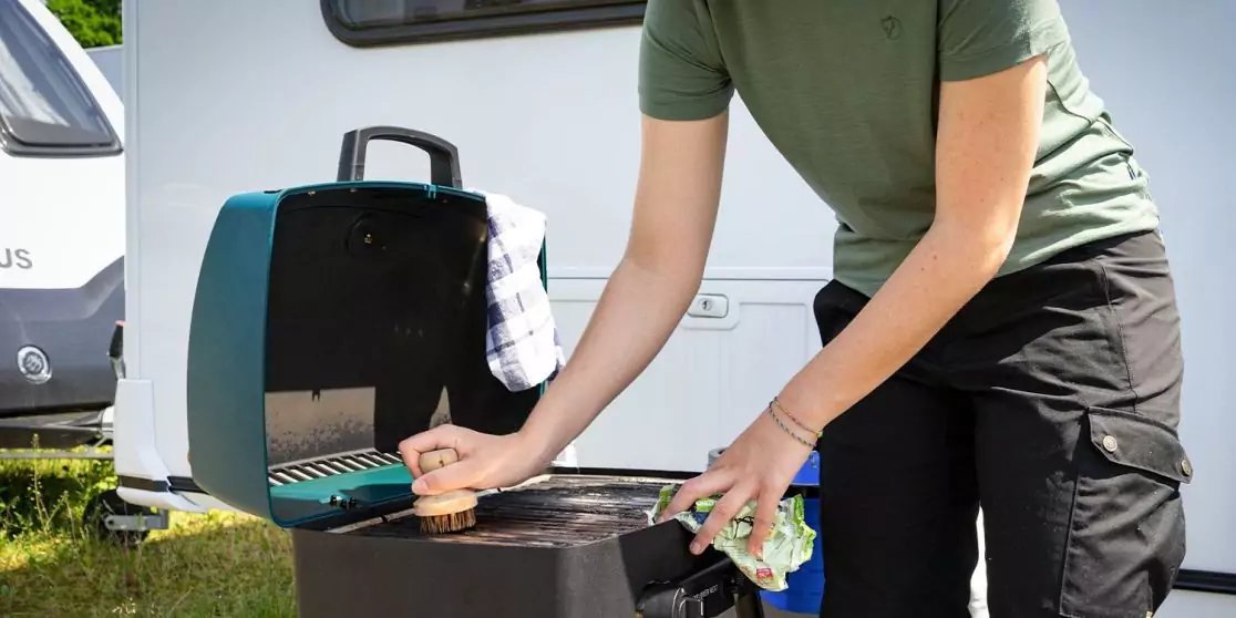 Person mit gr&uuml;nem T‑Shirt und schwarzer Hose neben ge&ouml;ffnetem tragbarem Grill vor einem Wohnwagen, legt mit linker Hand ein rundes Lebensmittel auf den Grillrost; blau‑wei&szlig;es Tuch am Deckel