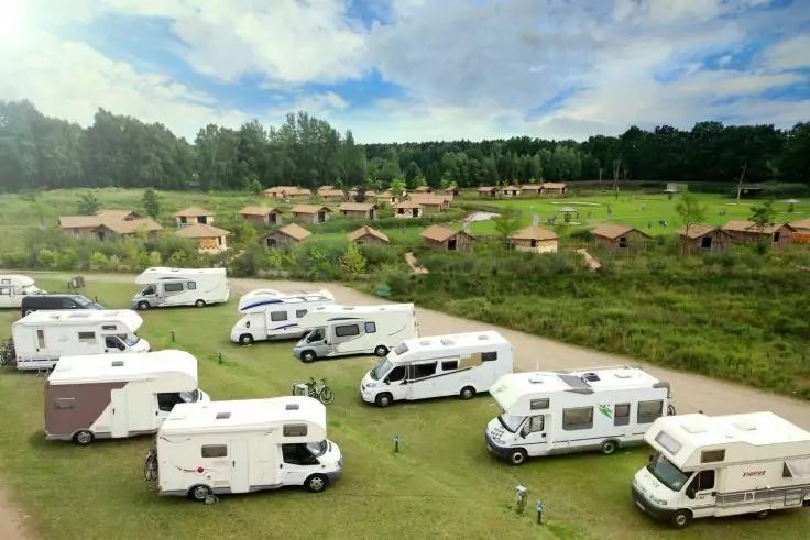Mehrere Wohnmobile und Wohnwagen auf einer Wiese im Vordergrund, dahinter Reihen kleiner Holzh&auml;user, B&auml;ume und bew&ouml;lkter Himmel