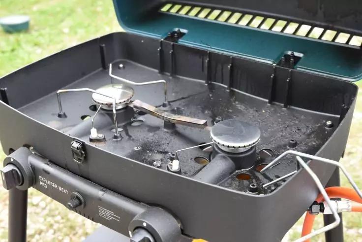 Schwarzer tragbarer Gasgrill mit offenem Deckel, zwei runden Brennern, verschmutzter Innenfl&auml;che und orangefarbenem Gasanschluss