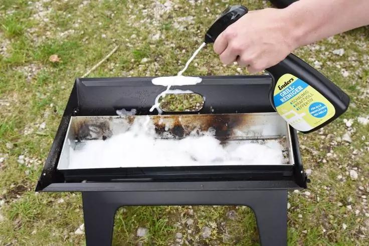 Hand gie&szlig;t Fl&uuml;ssigkeit aus schwarzer-gelber Plastikflasche auf wei&szlig;es faseriges Material in einer rechteckigen schwarzen Metallgrillwanne mit verkohlten R&auml;ndern, im Freien auf Gras
