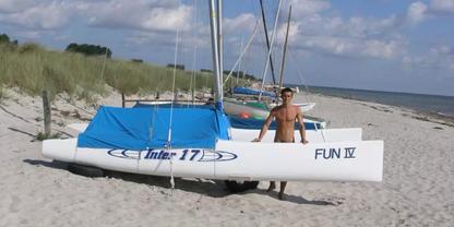 Sandstrand mit D&uuml;nen und mehreren kleinen Katamaran-Segelbooten mit blauen Abdeckungen; eine Person steht neben dem vordersten Boot mit der Aufschrift 'FUN IV'