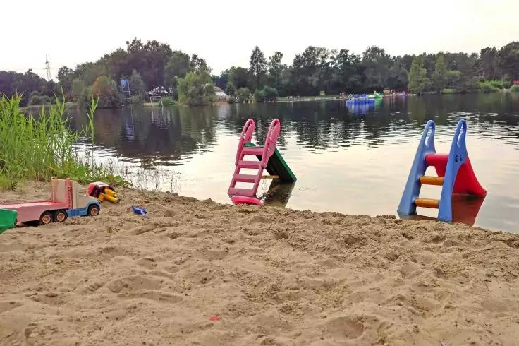 Sandstrand an einem See mit zwei bunten Plastikrutschen teilweise im flachen Wasser, Spielzeugwagen und Sandspielzeug am Ufer, B&auml;ume und H&auml;user im Hintergrund