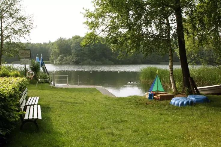 Ufer eines Sees mit Holzsteg und Leiter, zwei B&auml;nken links, Ruderboot rechts, blauem aufblasbarem Schwimmring und kleinem Spielzeugsegelboot auf dem Gras, B&auml;ume und Schilf im Hintergrund