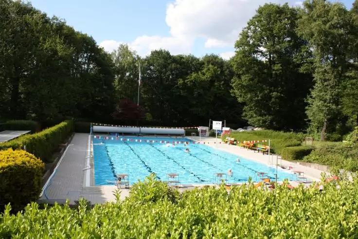 Au&szlig;en-Schwimmbecken mit markierten Bahnen, mehreren Schwimmern im Wasser, Beckenrand und B&auml;umen im Hintergrund