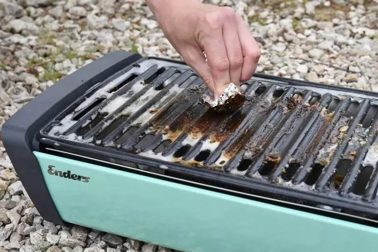 Hand h&auml;lt zusammengekn&uuml;lltes St&uuml;ck Aluminiumfolie &uuml;ber verschmutzten Grillrosten eines mintgr&uuml;nen Elektrogrills mit sichtbarem Schriftzug 'Enders', Ger&auml;t steht auf Kies