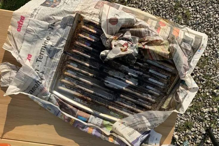 Metallgrillrost mit dunklen R&uuml;ckst&auml;nden auf zerknitterten Zeitungen, auf einem Holzbrett neben Kies