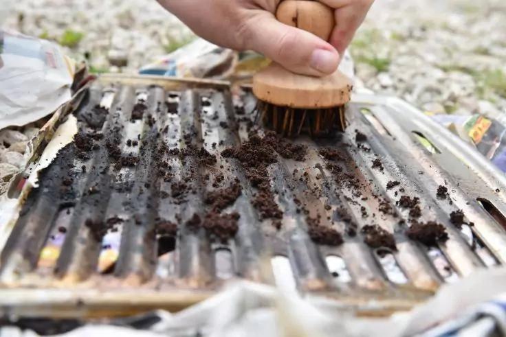 Hand h&auml;lt eine Holzb&uuml;rste und schrubbt ein Metallgrillrost mit dunklen Schmutzresten
