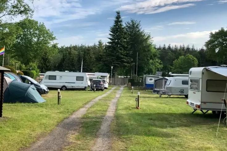 Campingplatz mit mehreren Wohnwagen und einem Zelt neben einer schmalen Fahrspur, Grasfl&auml;chen, B&auml;umen im Hintergrund und teils bew&ouml;lktem Himmel