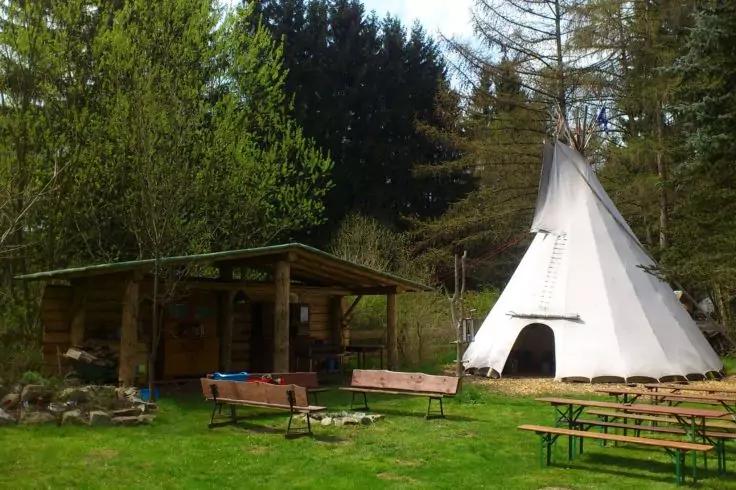 &Uuml;berdachter Holzunterstand links, wei&szlig;es Tipi rechts, B&auml;nke und Tische auf Rasen, Baumreihe im Hintergrund