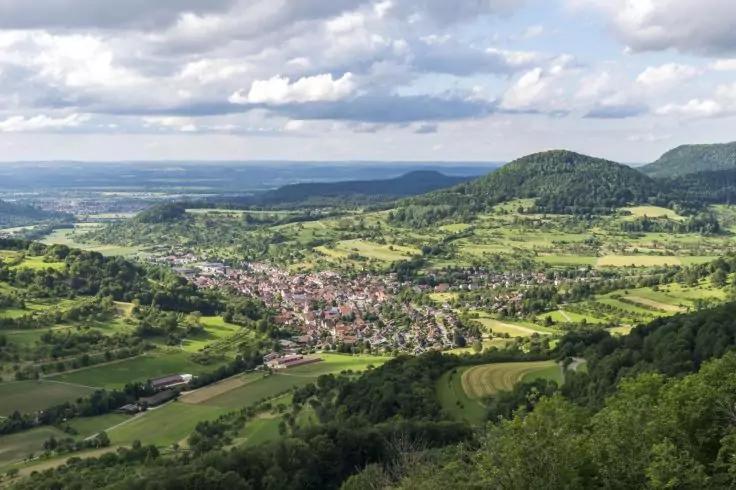Tal mit einer Ansammlung von Geb&auml;uden, umgeben von gr&uuml;nen Feldern, H&uuml;geln und bewaldeten H&auml;ngen, bew&ouml;lkter Himmel