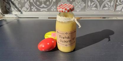 Glasflasche mit gelbem Inhalt, rot-wei&szlig; kariertem Deckel, Spitzenband und Papieretikett mit der Aufschrift "Frohe Ostern", daneben ein rotes Ei mit wei&szlig;en Punkten und ein gelbes Ei auf einer dunklen Arbeitsfl&auml;che vor gemusterten Fliesen