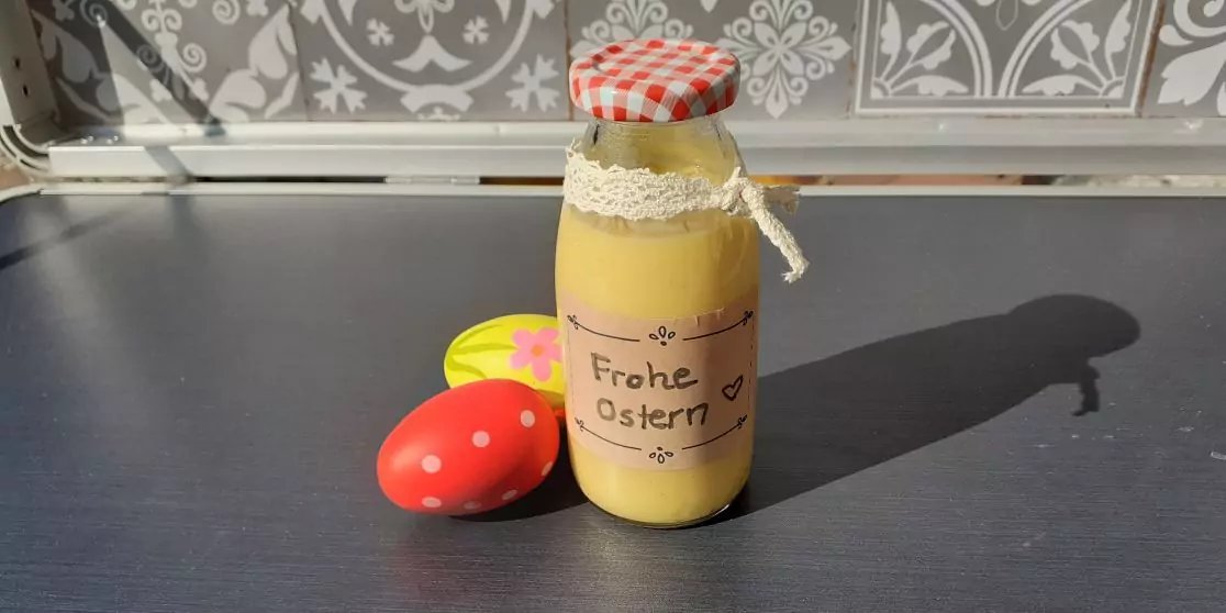 Glasflasche mit gelbem Inhalt, rot-wei&szlig; kariertem Deckel, Spitzenband und Papieretikett mit der Aufschrift "Frohe Ostern", daneben ein rotes Ei mit wei&szlig;en Punkten und ein gelbes Ei auf einer dunklen Arbeitsfl&auml;che vor gemusterten Fliesen