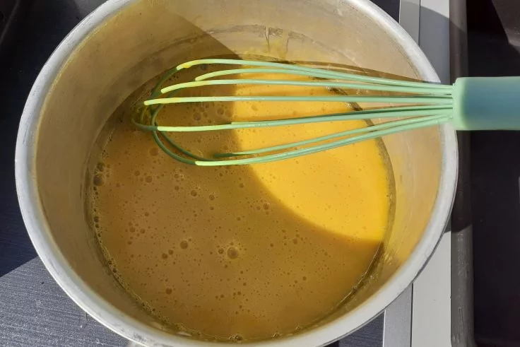 Metalltopf mit gelber Mischung und gr&uuml;nem Schneebesen auf Herdplatte