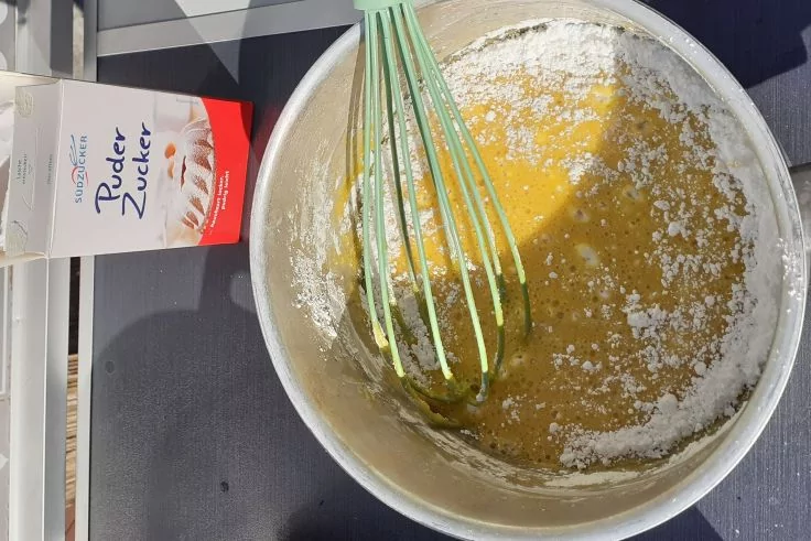 Metallr&uuml;hrsch&uuml;ssel mit gelber Mischung und wei&szlig;em Puderzucker am Rand, gr&uuml;ner Schneebesen darin, links Packung mit der Aufschrift 'Puderzucker'