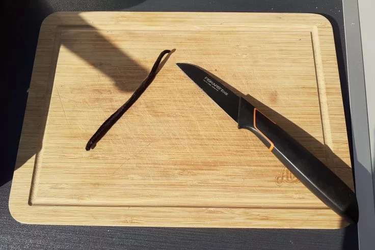 Rechteckiges Holzschneidebrett auf dunkler Unterlage, eine Vanilleschote und ein Messer mit schwarzem Griff; Sonnenlicht wirft Schatten