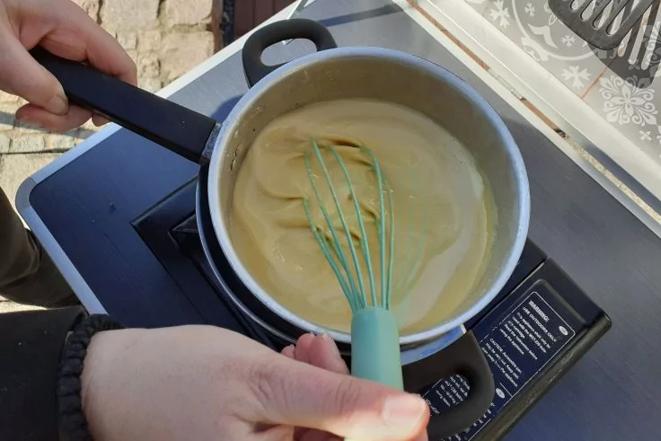 Zwei H&auml;nde r&uuml;hren mit einem hellblauen Schneebesen eine gelbe Mischung in einem Edelstahltopf auf einem tragbaren Campingkocher
