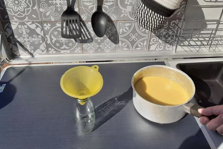 Wei&szlig;e Stielpfanne mit gelber Fl&uuml;ssigkeit, Hand am Griff, gelber Trichter in Glasflasche auf dunkler Arbeitsplatte, gemusterte Fliesen, h&auml;ngende K&uuml;chenutensilien und Abtropfgestell