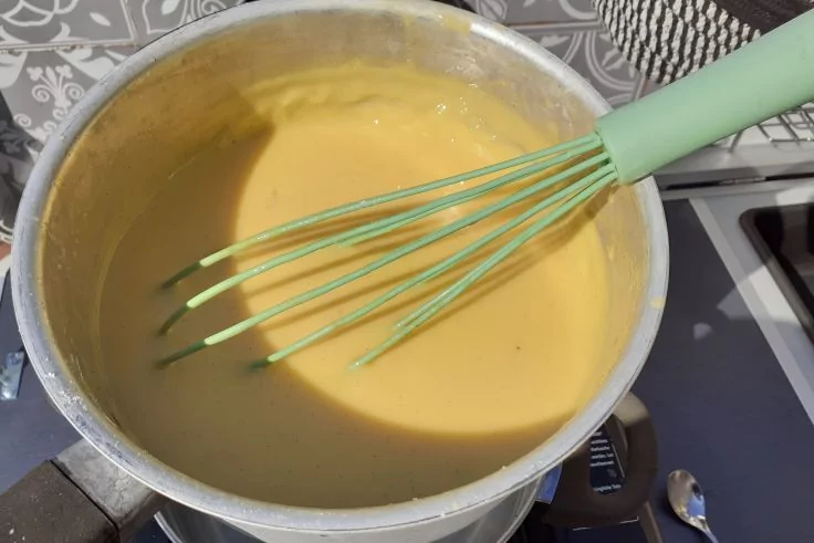 Metalltopf mit gelber Masse und hellgr&uuml;nem Schneebesen auf einem Herd; Topfgriff, L&ouml;ffel und gemustertes Tuch im Hintergrund sichtbar