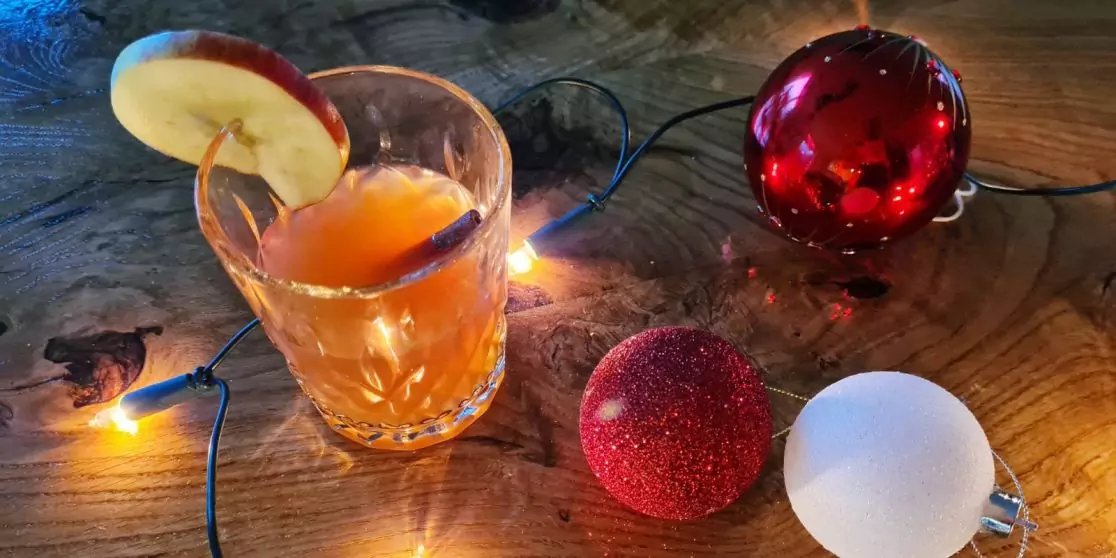 Glas mit orangefarbener Fl&uuml;ssigkeit und Apfelscheibe am Rand auf einer Holzoberfl&auml;che, Lichterkette mit warmwei&szlig;en L&auml;mpchen und drei Christbaumkugeln (rote gl&auml;nzende, rote glitzernde, wei&szlig;e)
