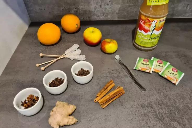 Grauer Tisch mit Orangen, kleinen &Auml;pfeln, einer Flasche mit gr&uuml;nem Etikett, drei Teebeuteln, drei wei&szlig;en Sch&auml;lchen mit dunklen Gew&uuml;rzen, einem Ingwerst&uuml;ck, Zimtstangen, einem Metalll&ouml;ffel und kleinen Holzst&auml;bchen