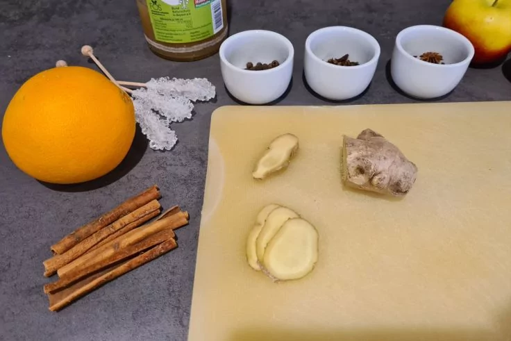 Orange, Zimtstangen, Kandiszucker auf Stiel, drei wei&szlig;e Schalen mit Gew&uuml;rzst&uuml;cken, Glas mit gr&uuml;nem Etikett, Apfel, Ingwer und Ingwerscheiben auf hellem Schneidebrett
