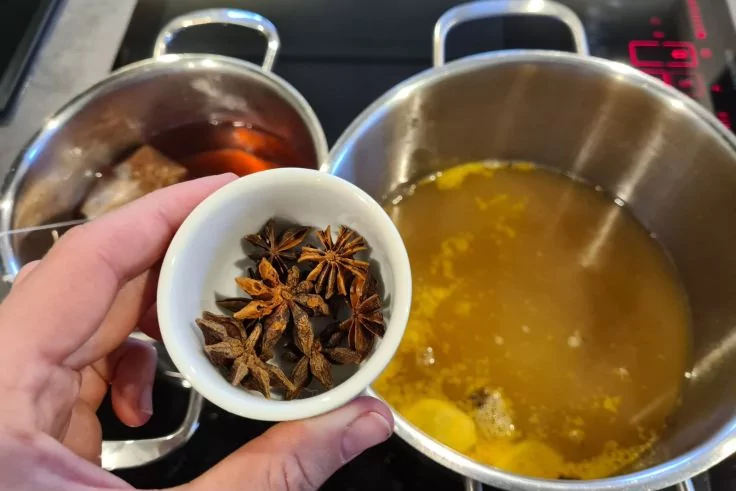 Hand h&auml;lt kleine wei&szlig;e Schale mit Sternanis &uuml;ber zwei Edelstahlt&ouml;pfen auf Kochfeld; linker Topf dunkle Fl&uuml;ssigkeit, rechter Topf gelbe Fl&uuml;ssigkeit mit Scheiben