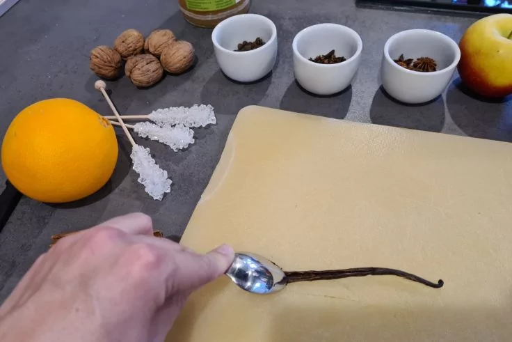 Hand mit L&ouml;ffel kratzt Mark aus einer aufgeschnittenen Vanilleschote, die auf einem hellen Teigblatt liegt; daneben Orange, Zuckerst&auml;bchen, Waln&uuml;sse, drei kleine wei&szlig;e Schalen mit braunen Gew&uuml;rzen und ein Apfel auf der Arbeitsfl&auml;che