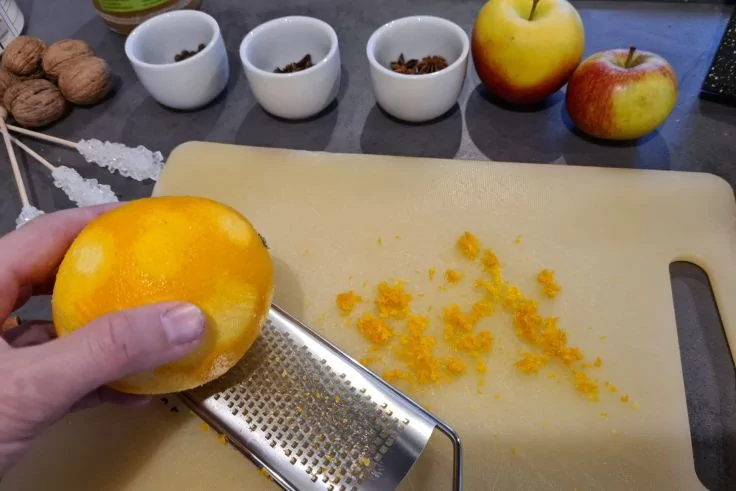 Hand reibt Orangenschale &uuml;ber einer feinen Reibe; Orangenzesten auf einem Schneidebrett, zwei &Auml;pfel, drei kleine wei&szlig;e Schalen mit braunen Gew&uuml;rzen und mehrere Waln&uuml;sse im Hintergrund