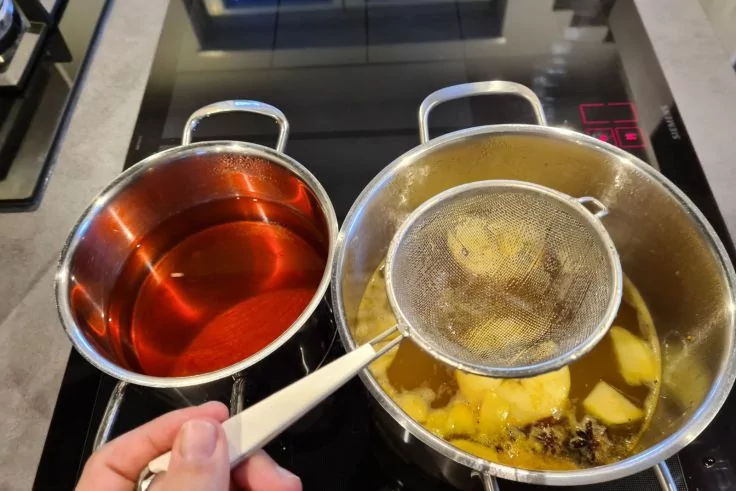 Zwei T&ouml;pfe auf schwarzem Ceranfeld: linker Topf mit dunkelroter Fl&uuml;ssigkeit, rechter Topf mit gelber Fl&uuml;ssigkeit und hellen Scheiben. Hand h&auml;lt ein feines Metallsieb &uuml;ber dem rechten Topf. Rotes Display am Herd sichtbar